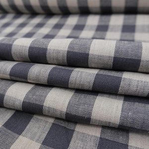 May include: A folded length of fabric featuring a classic gingham pattern with alternating squares of dark charcoal gray and natural beige. The textile displays a visible woven texture with a matte finish, arranged in multiple soft, horizontal folds.