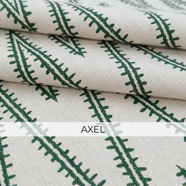 May include: A folded piece of off-white fabric featuring a repeating pattern of dark green, fern-like leaf stems arranged in diagonal lines. The word AXEL is printed in black capital letters across the center of the fabric surface.