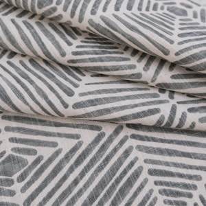 May include: A piece of fabric featuring a repeating geometric pattern of dark gray lines on an off-white background. The design consists of hexagonal shapes formed by parallel lines, with the fabric folded to show depth and texture.
