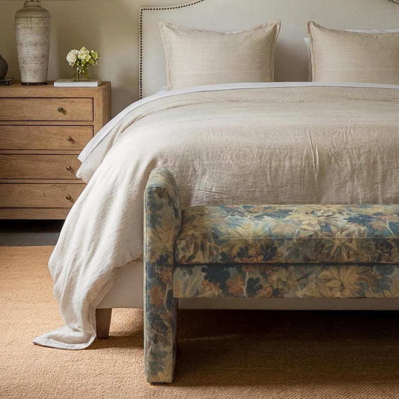 May include: A bedroom scene featuring a bed with a cream-colored duvet and pillows. A floral patterned bench sits at the foot of the bed. A wooden nightstand and a vase are visible in the background. The room has a neutral color palette.
