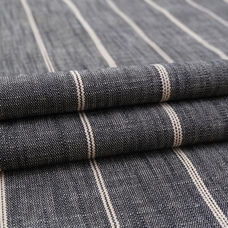 May include: Close-up of a folded fabric with a dark gray background and vertical white stripes. The fabric has a textured appearance, with the stripes evenly spaced across the material. The folds create shadows, adding depth to the image. This textile is suitable for various home decor projects.
