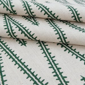 May include: A piece of off-white fabric featuring a repeating pattern of dark green, fern-like botanical lines arranged in diagonal rows. The textile has a visible woven texture and is folded to show the depth and drape of the material.