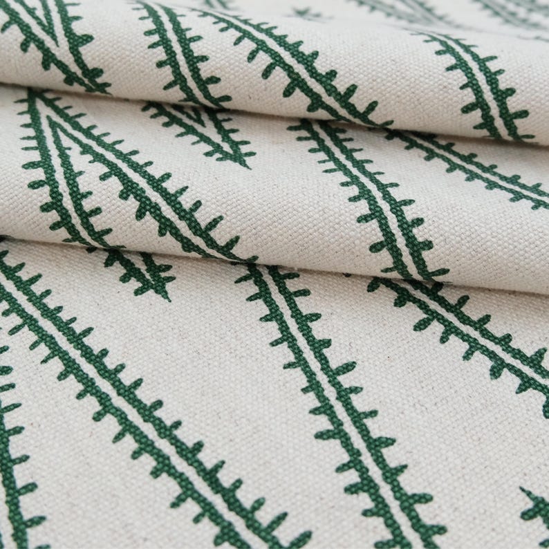 May include: A piece of off-white fabric featuring a repeating pattern of dark green, fern-like botanical lines arranged in diagonal rows. The textile has a visible woven texture and is folded to show the depth and drape of the material.