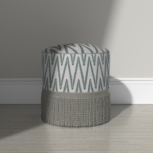 May include: A cylindrical ottoman featuring a light gray fabric top and sides with a dark green zigzag pattern. The base of the ottoman is adorned with a wide, textured gray fringe trim that encircles the entire bottom edge of the furniture piece.