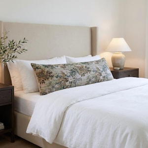 May include: A long rectangular bolster pillow featuring a detailed pastoral landscape print with muted green, brown, and beige tones. The pillow is positioned horizontally across a bed with white bedding, a beige headboard, and a bedside lamp.