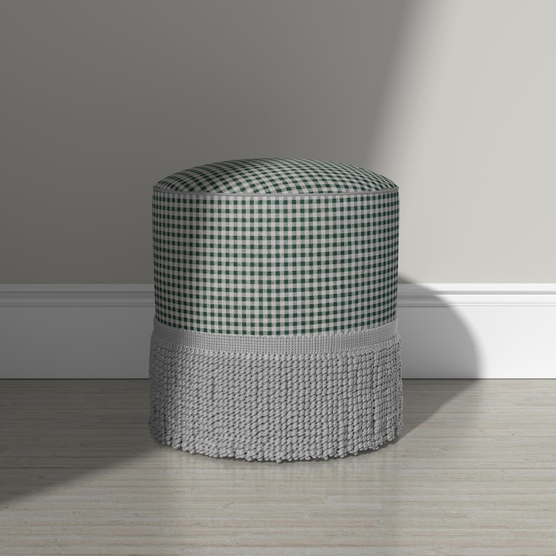 May include: A cylindrical ottoman upholstered in a dark green and white gingham check fabric. The base of the ottoman is adorned with a thick, light gray decorative fringe trim that encircles the bottom edge, providing a textured finish.