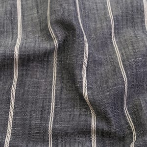 May include: Close-up of a textured fabric with vertical stripes. The fabric is a dark blue-gray color with thin, off-white stripes running from top to bottom. The fabric has a slightly wrinkled appearance.