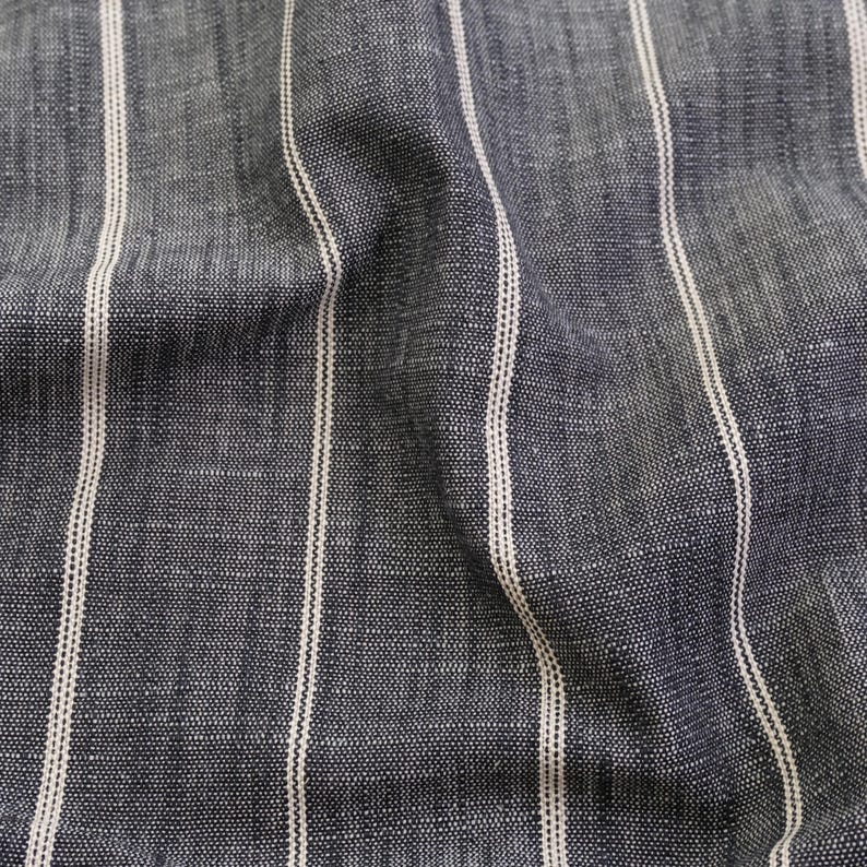 May include: Close-up of a textured fabric with vertical stripes. The fabric is a dark blue-gray color with thin, off-white stripes running from top to bottom. The fabric has a slightly wrinkled appearance.