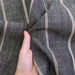 May include: Close-up of a dark gray fabric with vertical cream-colored stripes. The fabric is textured and appears to be woven. The material is being held, showing the folds and texture of the cloth.