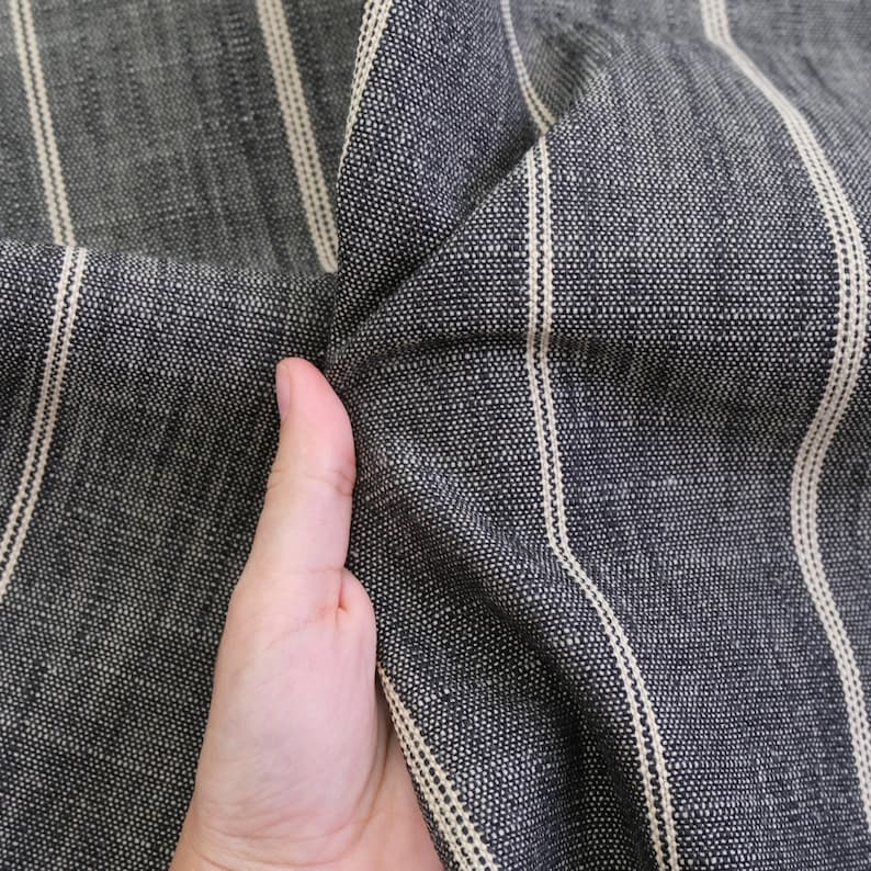 May include: Close-up of a dark gray fabric with vertical cream-colored stripes. The fabric is textured and appears to be woven. The material is being held, showing the folds and texture of the cloth.