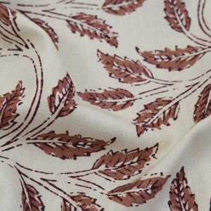 May include: A cream-colored fabric featuring a repeating pattern of brown leaves and stems. The textile has a visible woven texture with soft folds that create depth, showcasing the printed botanical design across the surface of the material.