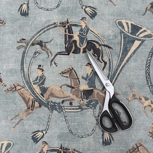 Puede incluir: Tijeras con mangos negros y plateados sobre una tela azul claro. La tela presenta un patr&oacute;n repetido de figuras ecuestres, caballos, perros y una trompa de caza en tonos marrones y azules. Borlas y dise&ntilde;os de cuerda completan el patr&oacute;n.
