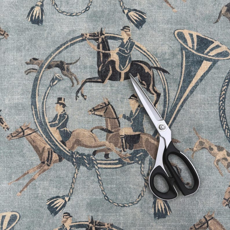 Puede incluir: Tijeras con mangos negros y plateados sobre una tela azul claro. La tela presenta un patr&oacute;n repetido de figuras ecuestres, caballos, perros y una trompa de caza en tonos marrones y azules. Borlas y dise&ntilde;os de cuerda completan el patr&oacute;n.
