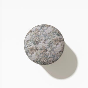May include: A round stool featuring a top upholstered in a fabric with a detailed botanical print in shades of green, brown, and cream. The circular seat has a smooth, flat surface and a thin, neutral-colored border around the outer edge.