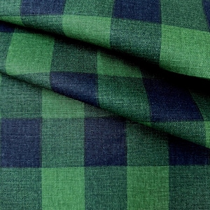 May include: Close-up of a green and navy blue plaid fabric. The fabric is folded, showing the texture and pattern of the woven material. The plaid design consists of large, evenly spaced squares.