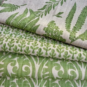 May include: A stack of three folded fabric pieces featuring a woven texture. The top layer displays a green fern leaf pattern on a light beige background, the middle layer has a small repeating green leaf motif, and the bottom layer shows a green scroll pattern.