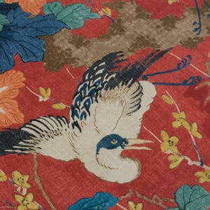 May include: A red fabric featuring a repeating pattern of white cranes with black and blue wing details, surrounded by orange and blue floral motifs, green leaves, and small yellow blossoms on thin brown branches across the textile surface.