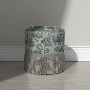 May include: A cylindrical ottoman featuring a light gray fabric upholstery with a dark green toile de jouy pattern depicting pastoral scenes. The base of the ottoman is adorned with a thick, textured gray fringe trim that encircles the bottom.