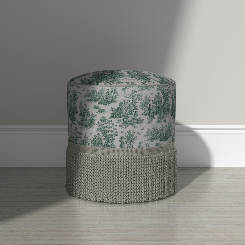 May include: A cylindrical ottoman featuring a light gray fabric upholstery with a dark green toile de jouy pattern depicting pastoral scenes. The base of the ottoman is adorned with a thick, textured gray fringe trim that encircles the bottom.