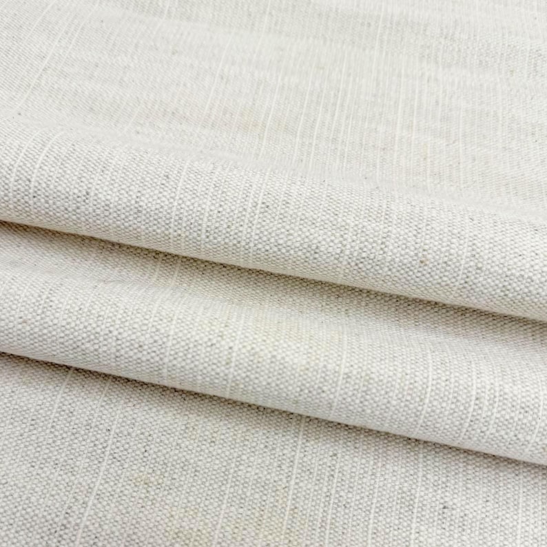 May include: A light beige fabric with a visible woven texture and subtle vertical stripes. The material features a natural, speckled appearance with a consistent weave pattern throughout, showing folds that create soft shadows across the surface.