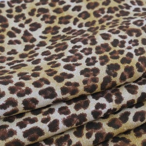 May include: A folded piece of fabric featuring a repeating leopard print pattern. The textile displays dark brown, irregular spots set against a light tan and beige background, creating a textured appearance with visible woven fibers throughout.