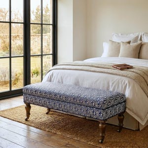 May include: A rectangular upholstered bench featuring a dark blue and white damask pattern, supported by four turned wooden legs with metal casters. The bench is positioned at the foot of a bed with white bedding and beige throw pillows.