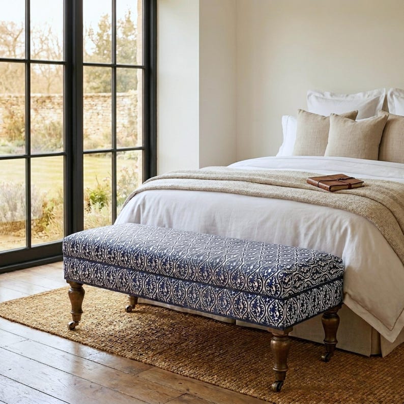 May include: A rectangular upholstered bench featuring a dark blue and white damask pattern, supported by four turned wooden legs with metal casters. The bench is positioned at the foot of a bed with white bedding and beige throw pillows.