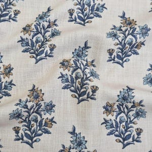 May include: A cream-colored fabric featuring a repeating pattern of floral sprigs in shades of blue and muted gold. The textile has a visible woven texture with subtle folds, showcasing a botanical design with leaves and blossoms arranged in clusters.