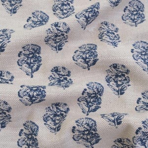 May include: A woven fabric featuring a repeating pattern of dark blue floral motifs printed on a light beige, textured background. The design consists of stylized, symmetrical flower shapes arranged in a consistent, evenly spaced grid layout.