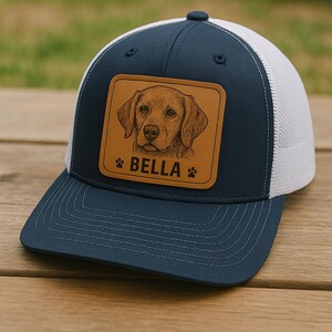 May include: A navy blue and white trucker hat with a brown leather patch featuring a dog portrait. The patch includes the name "BELLA" and paw prints. The hat has a curved brim and a mesh back.