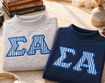 Könnte beinhalten: Ein hellgraues Sweatshirt und ein marineblaues Sweatshirt, beide mit den griechischen Buchstaben Sigma und Alpha in einem blau-weiß gestreiften Muster mit dickem blauem Rand, jeweils mit Stehkragen und langen Ärmeln versehen.