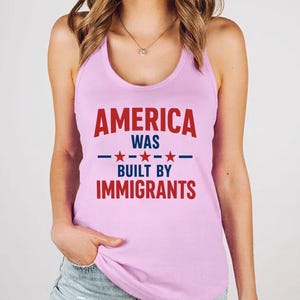 Pode incluir: Uma regata rosa claro com o texto "AMERICA WAS BUILT BY IMMIGRANTS" em vermelho e azul. O texto está centralizado na frente da regata, com uma linha azul e estrelas separando as palavras.