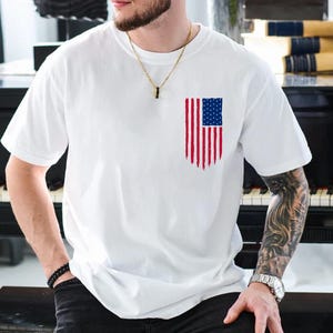 Pode incluir: Camiseta branca com um design desgastado da bandeira americana no peito. O gráfico da bandeira inclui listras vermelhas e brancas e um quadrado azul com estrelas brancas. A camiseta tem mangas curtas e gola redonda.