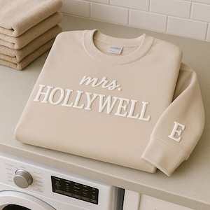 May include: Beige sweatshirt with the text "mrs. HOLLYWELL" in white, with the letter "E" on the sleeve. The sweatshirt is folded on a countertop next to a stack of beige towels.