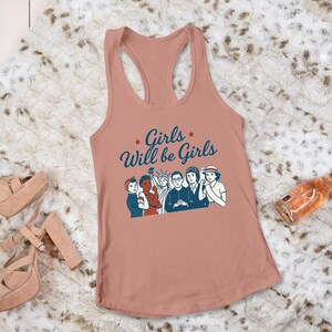 Pode incluir: Uma regata coral com o texto "Girls Will be Girls" e um gr&aacute;fico de mulheres. Uma garrafa de vinho ros&eacute; e saltos plataforma tamb&eacute;m s&atilde;o vis&iacute;veis.