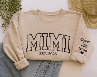 May include: A beige sweatshirt with the word "MIMI" in black block letters and "EST. 2025" underneath. The right sleeve has the names "Sophia, Gill, Olivia" and a heart. The sweatshirt is folded on blue jeans.