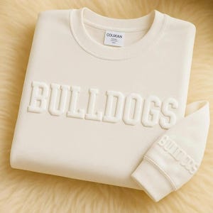 May include: Cream-colored sweatshirt with the word "BULLDOGS" in raised lettering across the front. The matching sleeve cuff also features the word "BULLDOGS". The sweatshirt has a crew neck and a small tag that reads "COUXIAN".