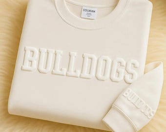 May include: Cream-colored sweatshirt with the word "BULLDOGS" in raised lettering across the front. The matching sleeve cuff also features the word "BULLDOGS". The sweatshirt has a crew neck and a small tag that reads "COUXIAN".