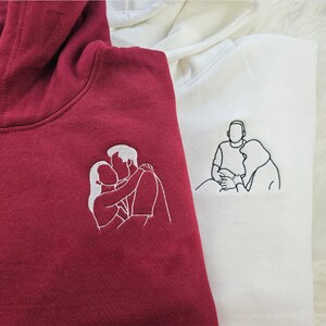 May include: Two hooded sweatshirts, one burgundy and one white, each embroidered with a line drawing of a couple. The burgundy hoodie features a couple kissing, while the white hoodie shows a couple embracing. Both hoodies have a drawstring hood.