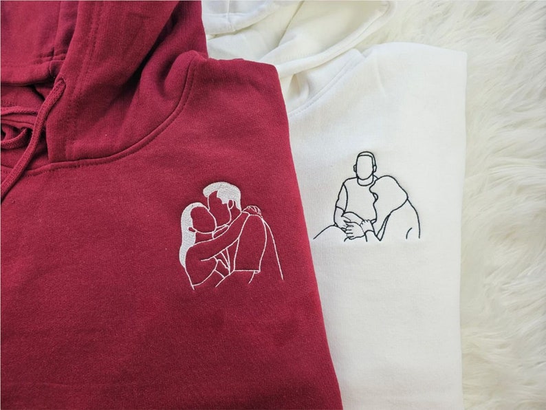 May include: Two hooded sweatshirts, one burgundy and one white, each embroidered with a line drawing of a couple. The burgundy hoodie features a couple kissing, while the white hoodie shows a couple embracing. Both hoodies have a drawstring hood.
