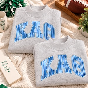 May include: Two light grey crewneck sweatshirts featuring the Greek letters Kappa Alpha Theta in a blue and white striped pattern. The letters are appliqued in a collegiate font across the chest of each garment, which has a ribbed collar.