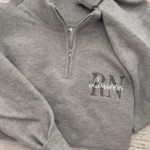 May include: A gray sweatshirt with a quarter-zip neckline and long sleeves. The embroidered text "Lauren RN" is visible on the front. The sweatshirt is folded, showing the texture of the fabric.
