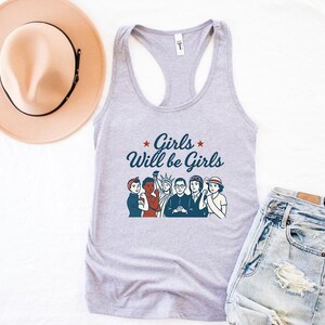 Pode incluir: Uma regata cinza claro com o texto "Girls Will be Girls" e uma ilustra&ccedil;&atilde;o de v&aacute;rias mulheres. Um chap&eacute;u bege com uma faixa preta e um par de jeans de lavagem clara tamb&eacute;m est&atilde;o na imagem.