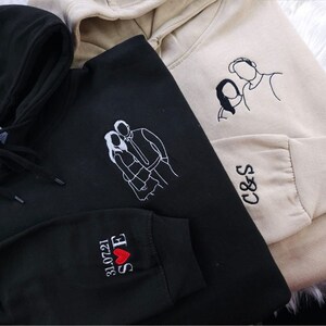 May include: Three hooded sweatshirts in black and beige. Each sweatshirt features embroidered line art of a couple. One black sweatshirt has the date 31.07.21, initials, and a heart on the sleeve. The other beige sweatshirt has initials on the sleeve.