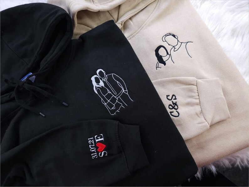 May include: Three hooded sweatshirts in black and beige. Each sweatshirt features embroidered line art of a couple. One black sweatshirt has the date 31.07.21, initials, and a heart on the sleeve. The other beige sweatshirt has initials on the sleeve.