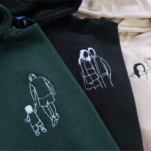 May include: Four hooded sweatshirts in dark green, black, and beige. Each sweatshirt features a white embroidered line drawing of a family or couple. The drawings include a father and child, a couple embracing, and a parent with a child.
