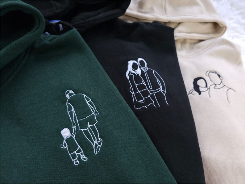 May include: Four hooded sweatshirts in dark green, black, and beige. Each sweatshirt features a white embroidered line drawing of a family or couple. The drawings include a father and child, a couple embracing, and a parent with a child.