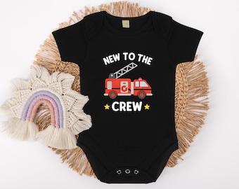 May include: Black baby bodysuit with the text "NEW TO THE CREW" and a red fire engine graphic. The bodysuit is short-sleeved and has a snap closure. A decorative rainbow and woven placemat are in the background.