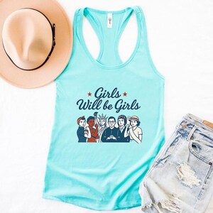 Pode incluir: Uma regata azul clara com o texto "Girls Will be Girls" e um gr&aacute;fico de mulheres. Um chap&eacute;u bege com uma faixa preta e cal&ccedil;&otilde;es de ganga tamb&eacute;m s&atilde;o vis&iacute;veis.