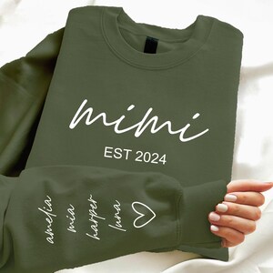 Könnte beinhalten: Ein grüner Pullover mit dem Text "mimi est 2024" auf der Vorderseite und den Namen "amelia", "mia", "harper", "luna" und einem Herz auf dem Ärmel.